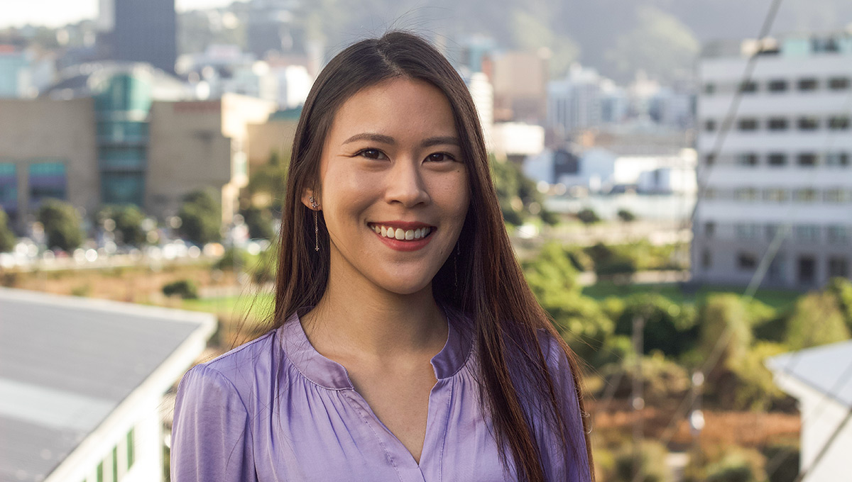 Portrait image of Ainsley Chin smiling in a purple blouse. Background is Wellington city including Te Papa and Waitangi Park