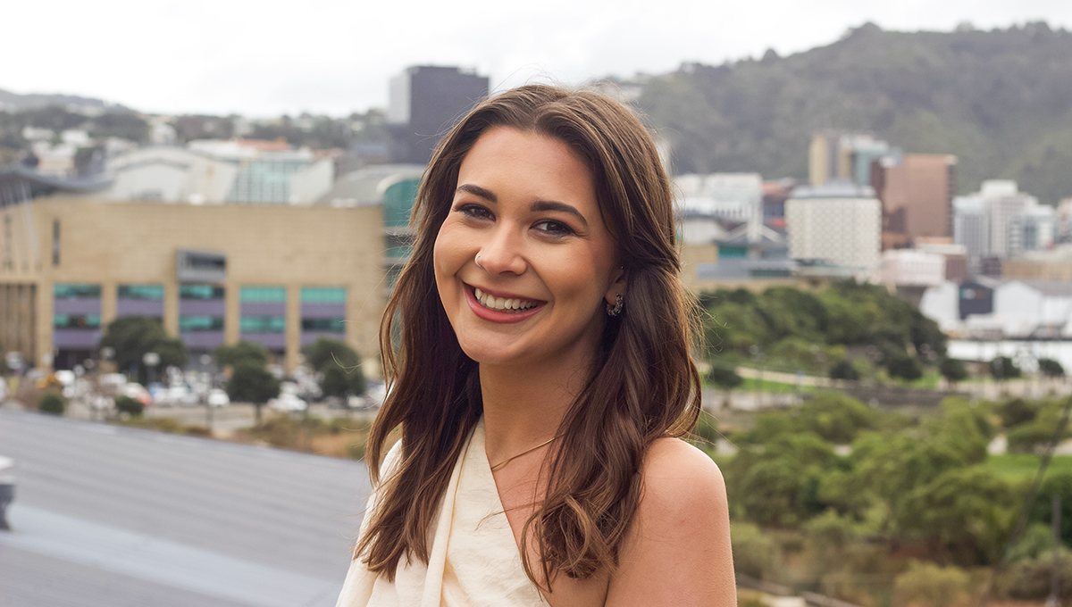 Landscape image of Ashely smiling. She is in a white off the shoulder long sleeve top. Her hair is brown. The background is of Wellington scenery with Waitangi park and Te Papa.